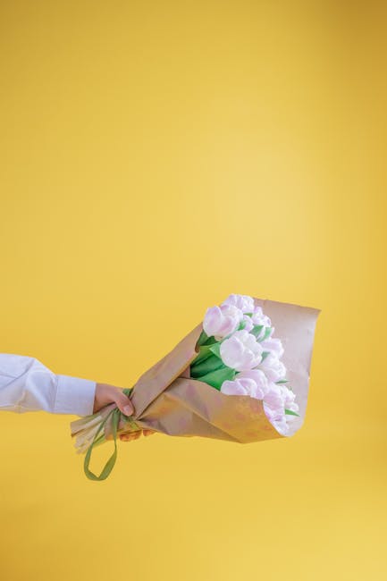 A hand, partially visible and dressed in a white shirt, holds a bouquet of soft pink tulips wrapped in brown paper with a green ribbon, set against a bright yellow background. The tulips have smooth p