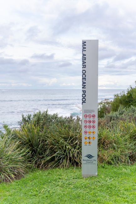 A tall, vertical white sign with black text reading 'Malabar Ocean Pool' stands amidst green shrubbery and grassy terrain near the coastline. The sign displays several symbols indicating rules or rest