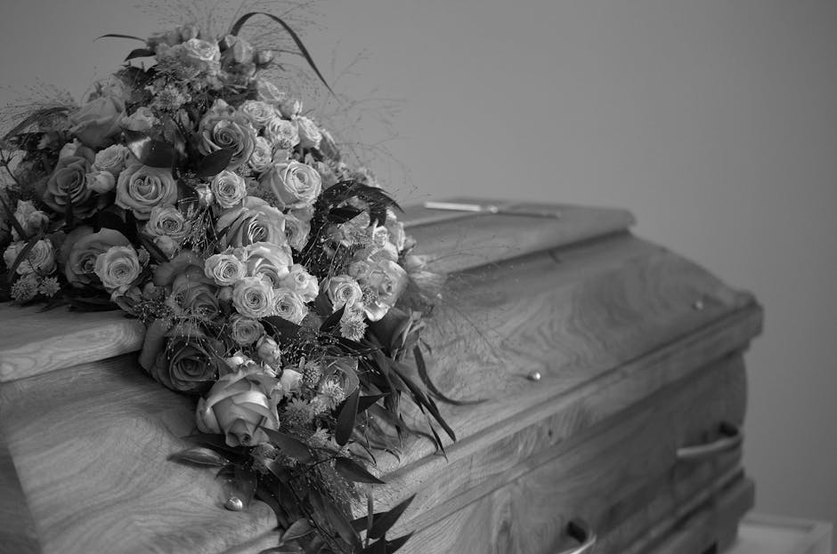 A large, elegant floral arrangement resting on the lid of a wooden casket, consisting of numerous roses in varying sizes, with some fully bloomed and others as buds, primarily in light shades, possibl