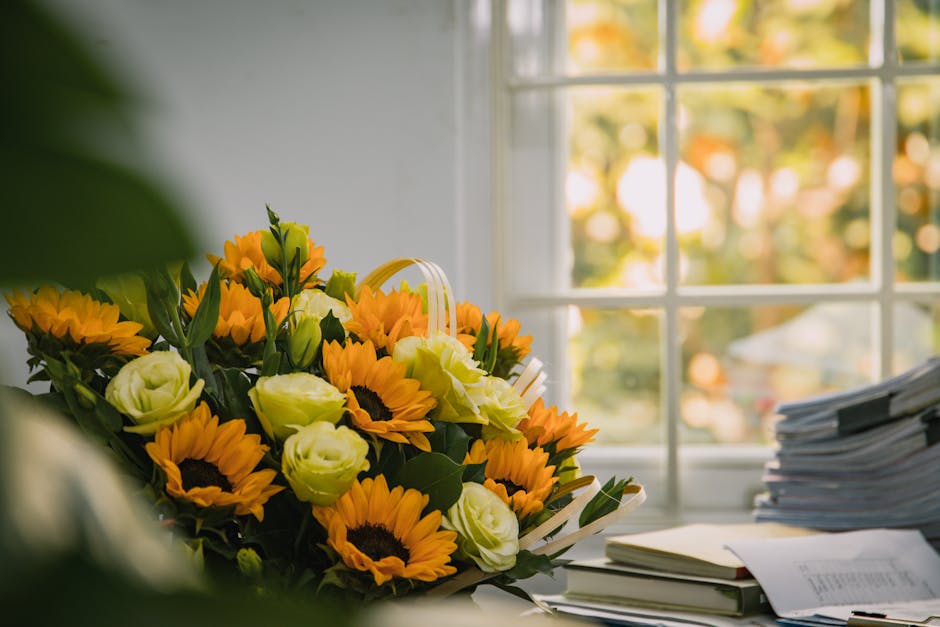 A vibrant bouquet of fresh flowers featuring large yellow sunflowers with dark centers, soft white roses, and green foliage arranged in a natural style. The bouquet is placed on a cluttered desk next
