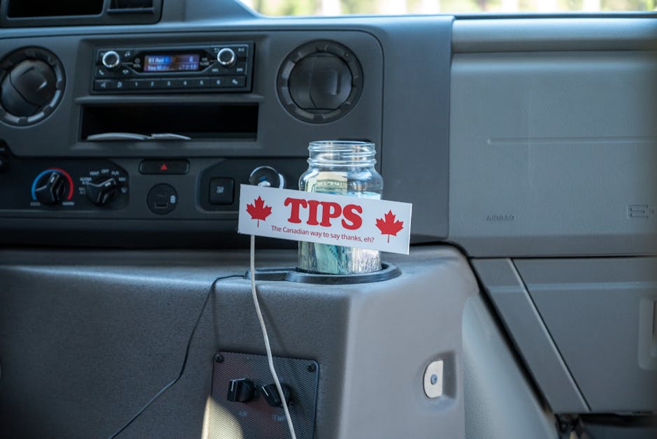 A clear glass jar containing assorted Canadian dollar bills positioned in the cupholder of a vehicle's dashboard, with a white card attached to the jar displaying the word 'TIPS' in bold red letters,