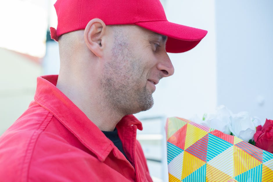 A man dressed in a red uniform, including a cap and jacket, is holding a colorful, geometric-patterned gift bag filled with white and red flowers. The arrangement features fresh, vibrant petals with v